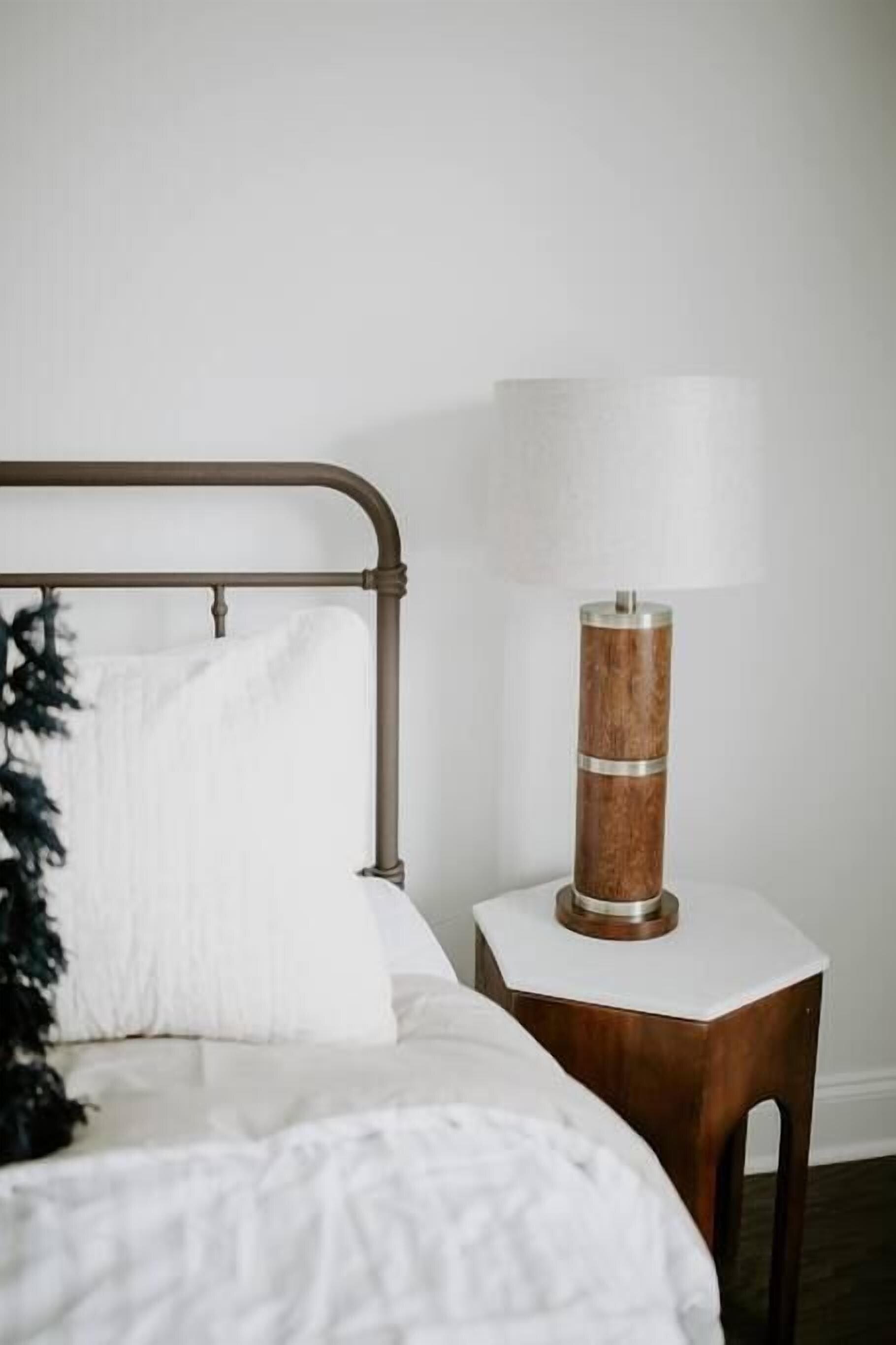 Una cama bien hecha con edredón blanco y una sola almohada blanca. Junto a la cama, una mesita de noche de madera con una lámpara cilíndrica.