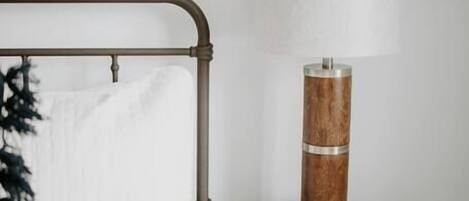 Una cama bien hecha con edredón blanco y una sola almohada blanca. Junto a la cama, una mesita de noche de madera con una lámpara cilíndrica.