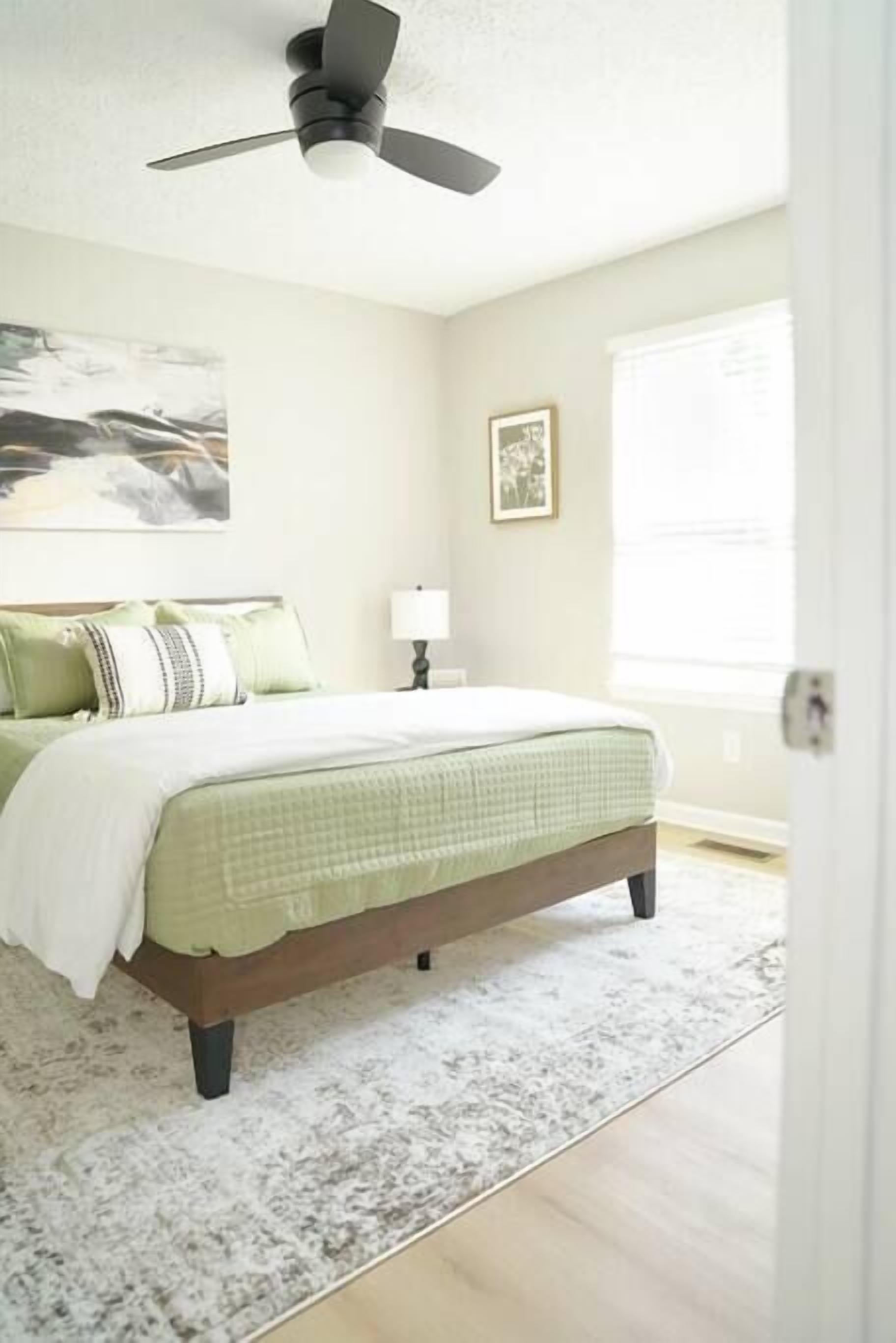 A bedroom with a bed, a ceiling fan, a nightstand, a lamp, a framed picture on the wall, and a window with blinds.