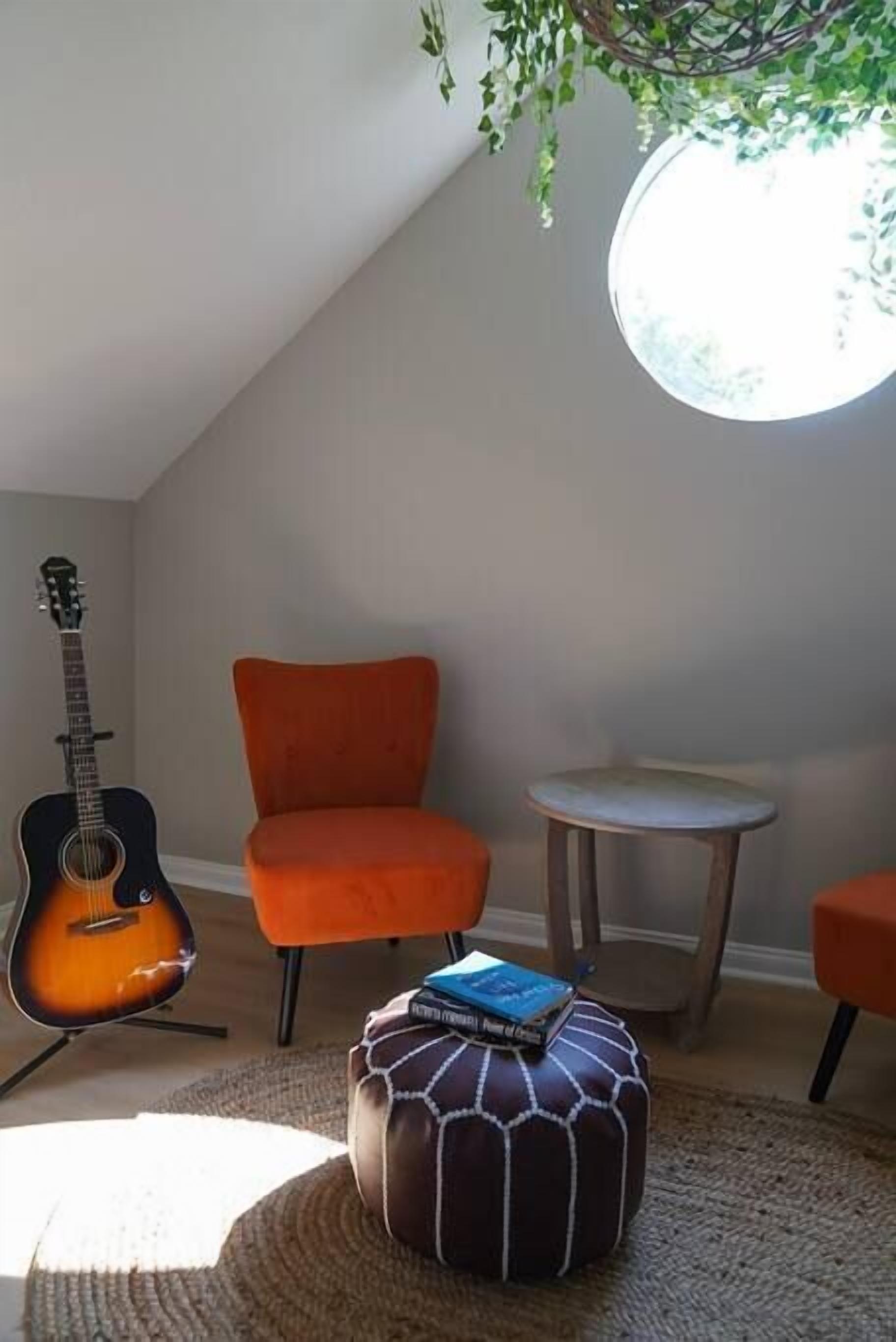 A room with a sloped ceiling, a guitar, an orange chair, a round table, and a small ottoman with books.