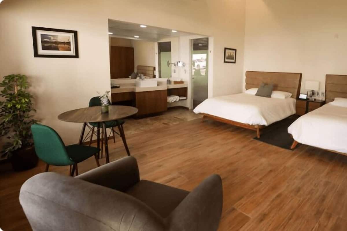 A hotel room with two beds, a sitting area with a chair and table, a bathroom with a mirror, and a potted plant.
