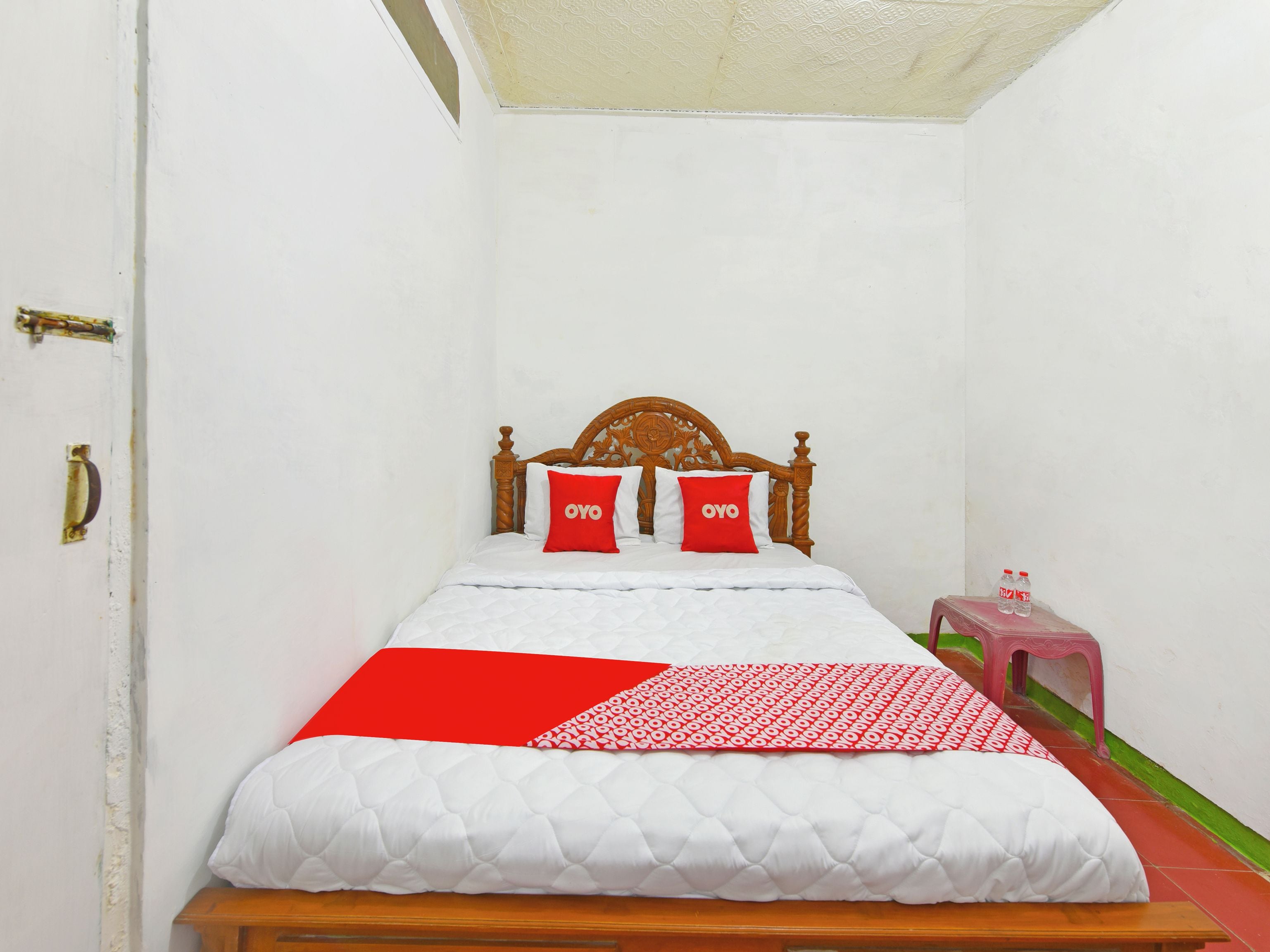 Habitación con una cama individual, cabecera de madera, dos almohadas rojas con la marca 'OYO', edredón blanco acolchado y manta con diseño rojo y blanco.