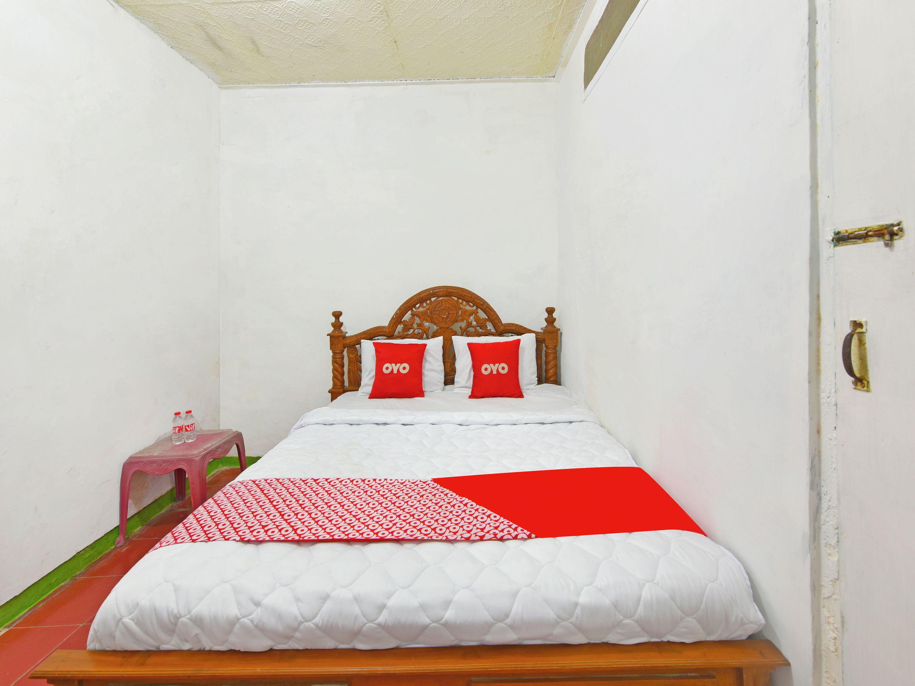 Uma cama de solteiro com cabeceira de madeira, duas almofadas vermelhas com a inscrição "OYO", um colchão acolchoado branco e um cobertor com padrão vermelho e branco.
