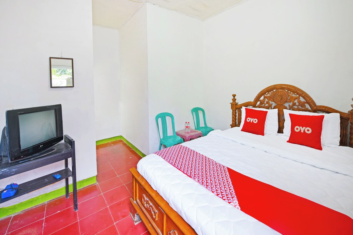 Habitación de hotel con una cama, un televisor sobre un soporte, una mesita con una silla y una colcha de edredón con estampado rojo y blanco.