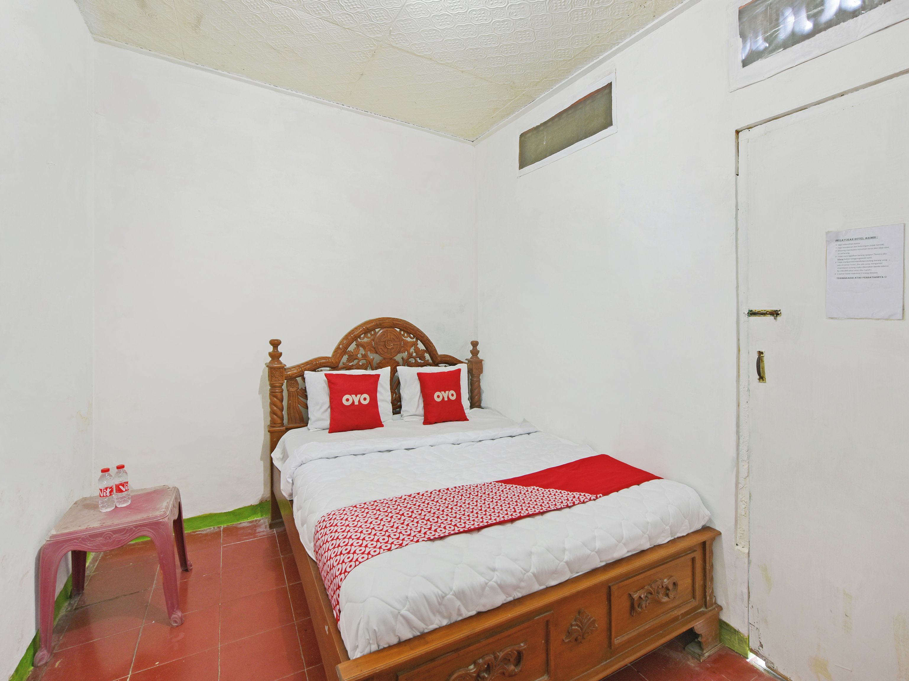 Habitación con una cama individual, cabecera de madera, dos almohadas rojas, una colcha blanca y una manta roja. Se observa un taburete de plástico con dos botellas encima, y una puerta con un letrero.