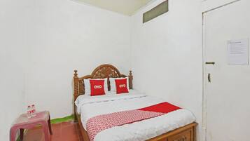 Habitación con una cama individual, cabecera de madera, dos almohadas rojas, una colcha blanca y una manta roja. Se observa un taburete de plástico con dos botellas encima, y una puerta con un letrero.