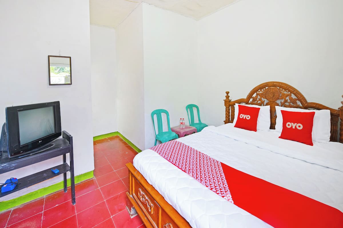 Habitación de hotel con una cama, un televisor sobre un soporte, una mesita con una silla y una colcha de edredón con estampado rojo y blanco.