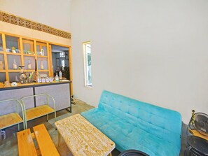 Una habitación con un sofá azul, mesas de madera, una zona de bar con frascos y botellas de vidrio, y una ventana.