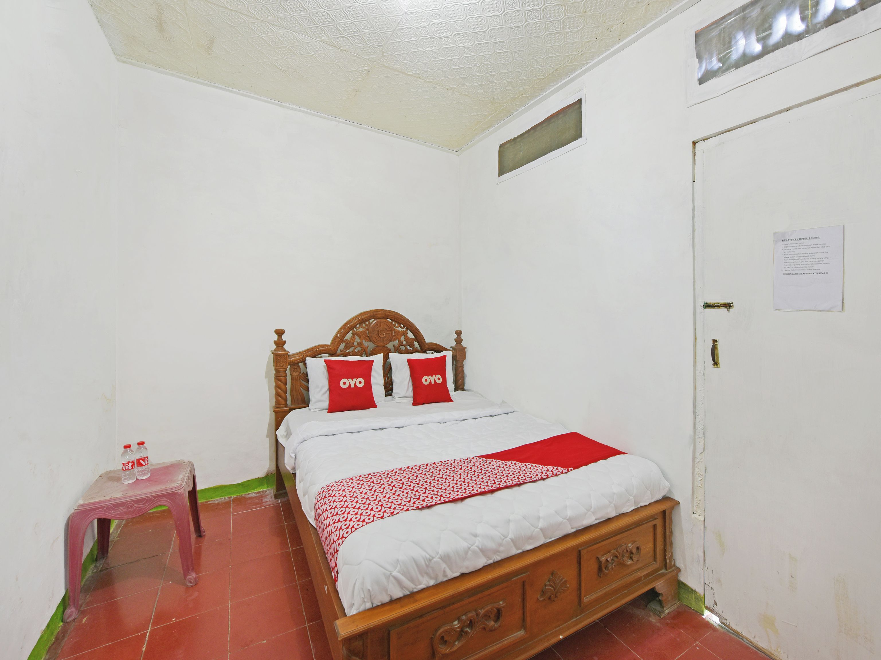 Quarto individual com uma cama de solteiro, cabeceira de madeira, roupa de cama branca e almofadas vermelhas. Um banquinho rosa pequeno com duas garrafas fica ao lado da cama.