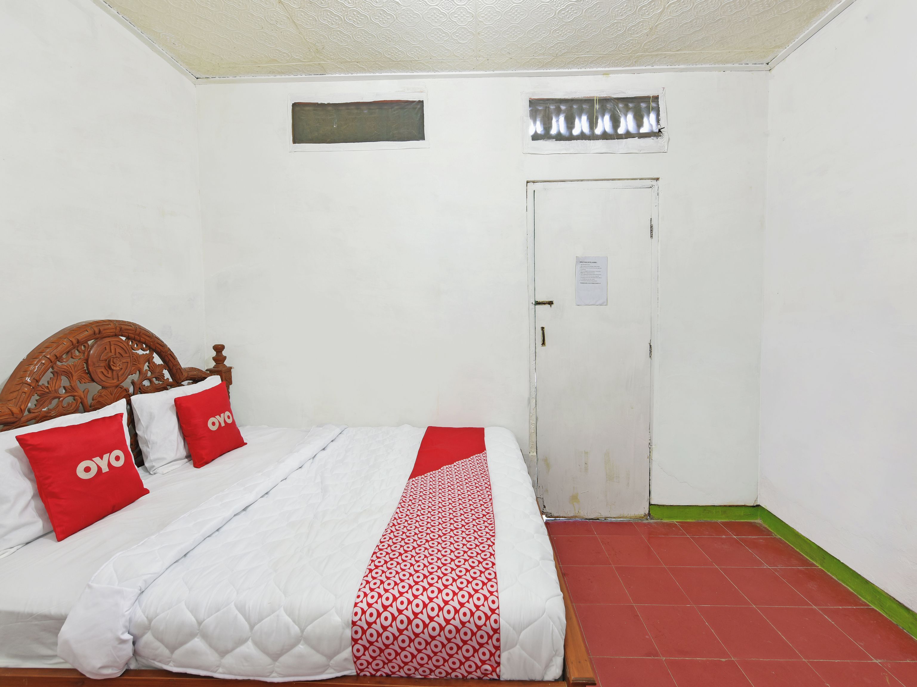 Una cama individual con cabecera de madera, ropa de cama blanca y almohadas rojas con el texto "oyo". Una manta roja y blanca estampada está colocada sobre la cama. La habitación tiene una puerta blanca con un letrero pequeño, una ventana con contraventanas de metal y un suelo de baldosas rojas.