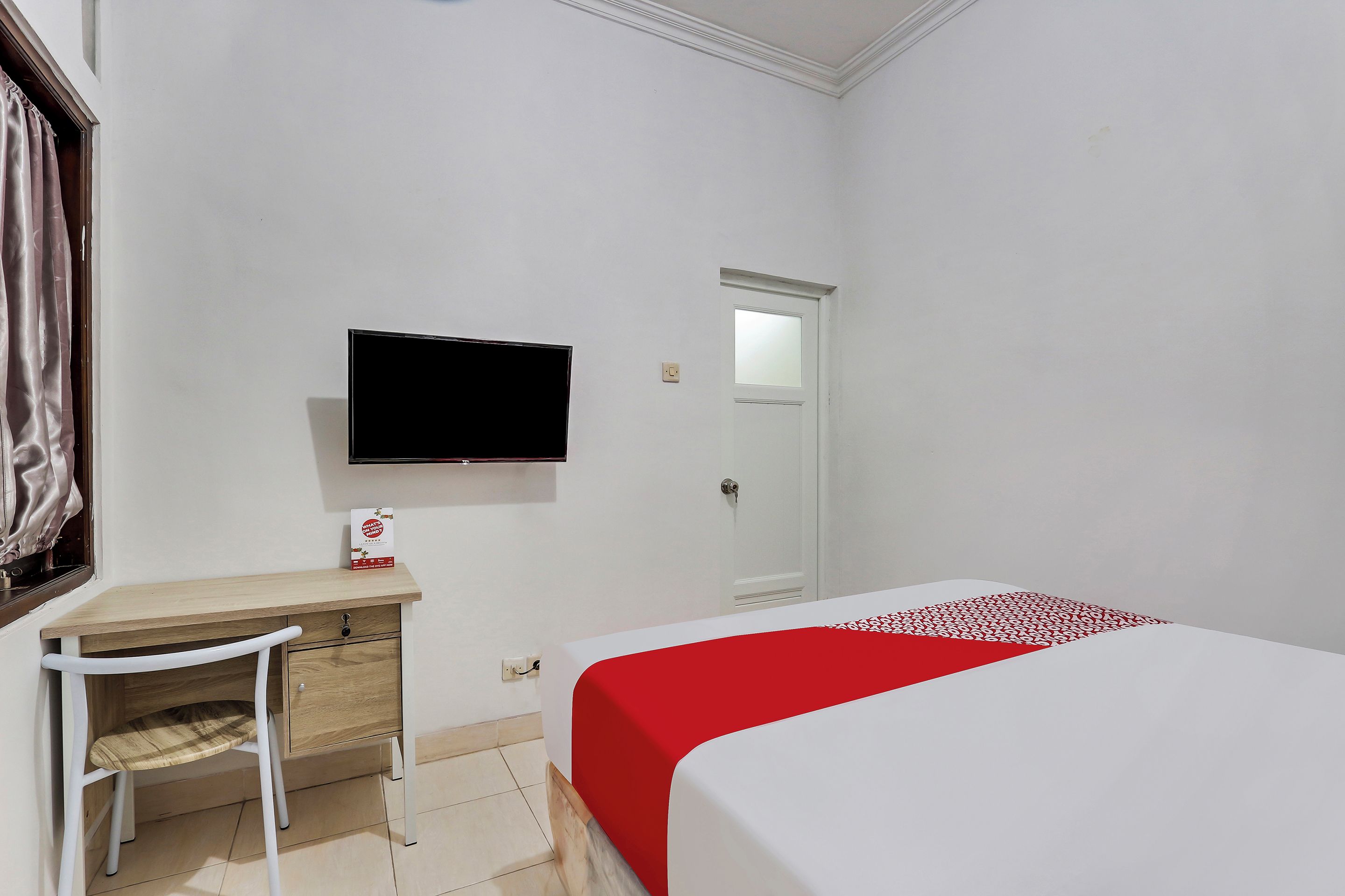 A hotel room with a bed, a desk with a chair, a television mounted on the wall, and a door.