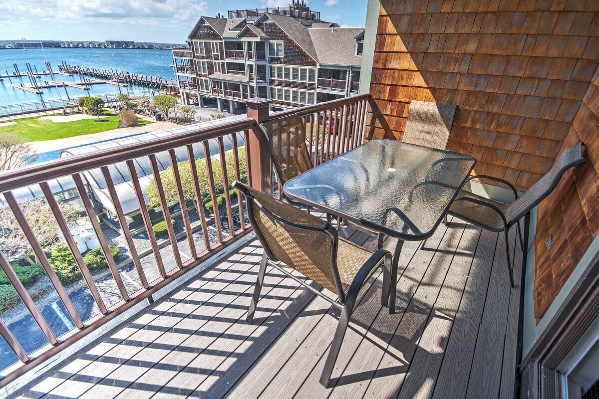 Een balkon met een glazen tafel en een rieten stoel, met uitzicht op een jachthaven en gebouwen.