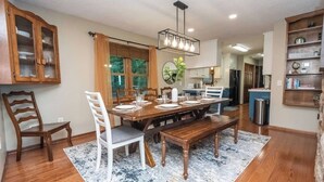 A dining area with a wooden table, white chairs, a chandelier, and a view of the kitchen. - Fall Colors | Prayer Mountain View Cabin | Fabulous Views from Fire Pit | Ping Pong (Moravian Falls)