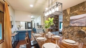 A dining area with a long wooden table set for four, a stone accent wall, and a kitchen in the background.