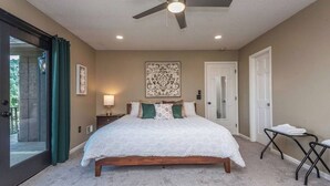 A bedroom with a large bed, a ceiling fan, a nightstand, a lamp, a door, and a window with curtains.