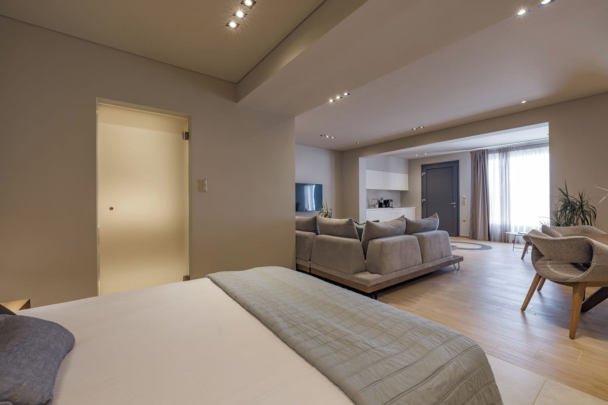 A modern hotel room with a large bed, a sofa, a dining area, and a kitchen in the background.