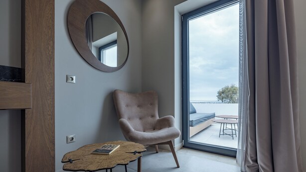 A modern room with a large window, a round mirror, a tufted armchair, and a small table with a book. - El mar Naxos  (Naxos)