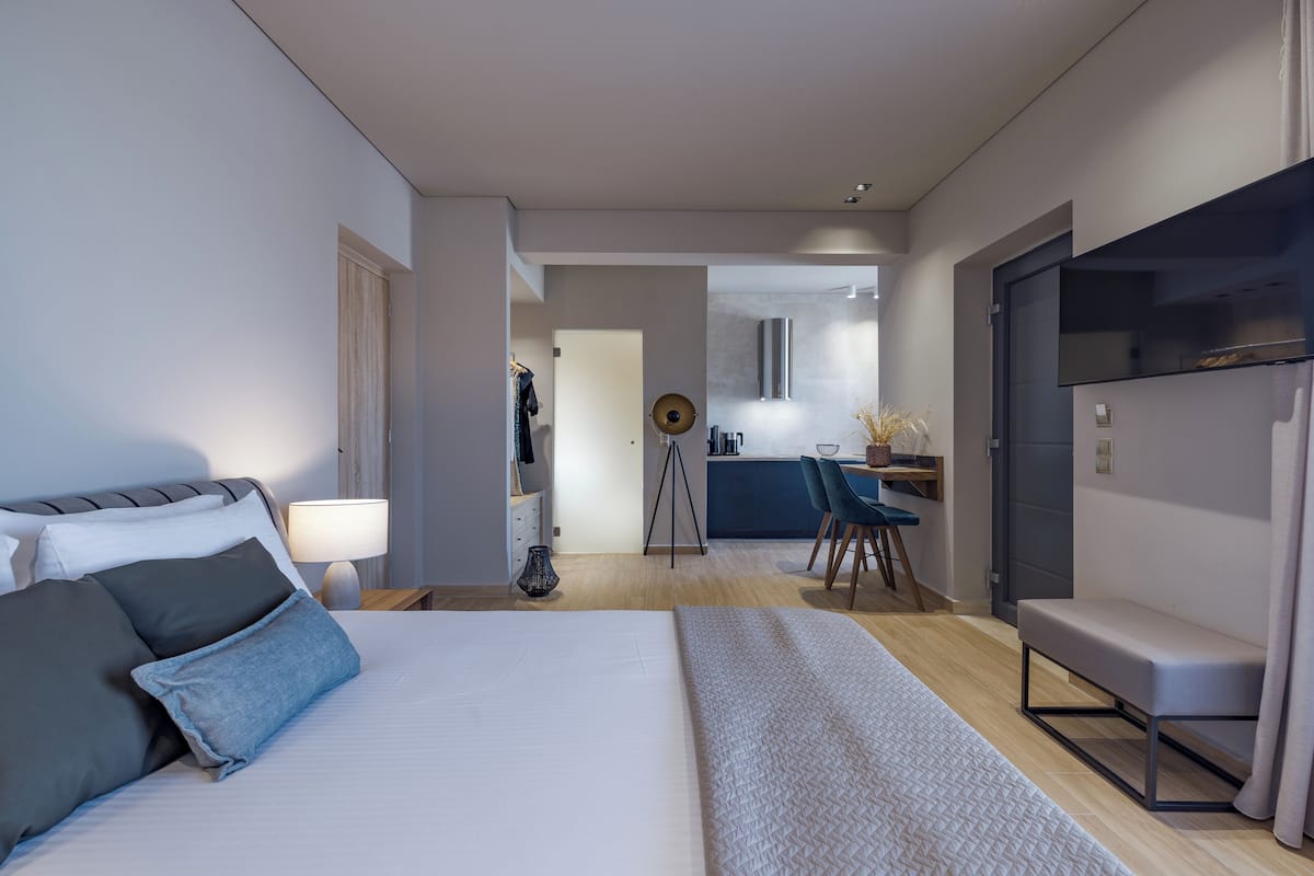 A modern hotel room with a large bed, a desk, and a kitchenette in the background.
