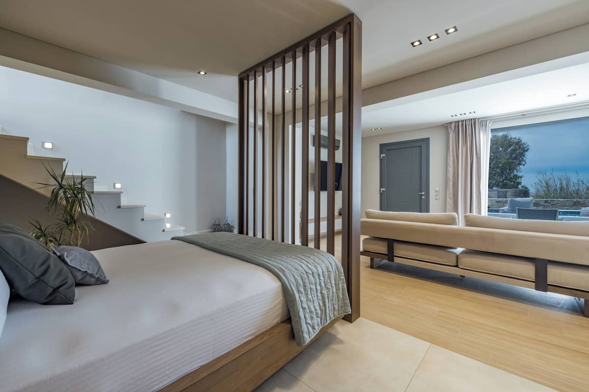 A modern bedroom with a large bed, a wooden partition, a sofa, and a view of the outdoors.