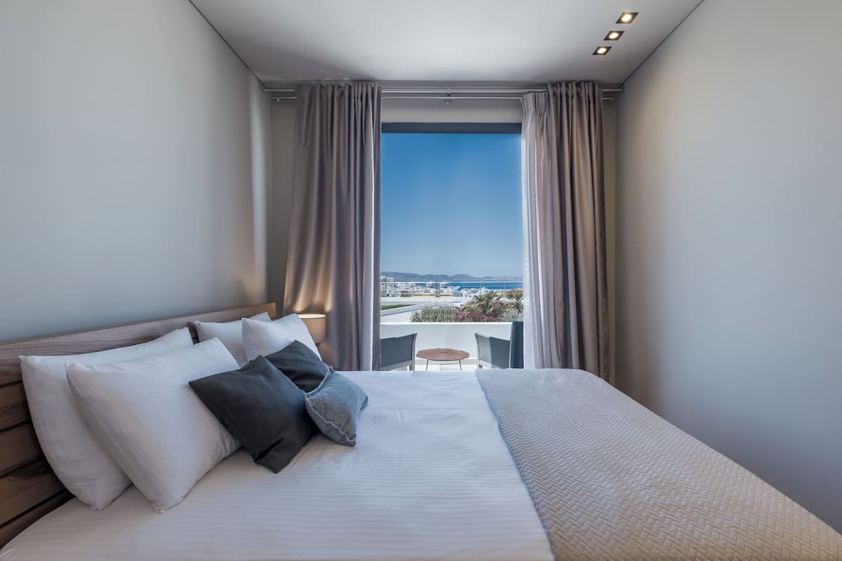 A modern hotel room with a large bed, a view of the city, and a balcony with a table and chairs.