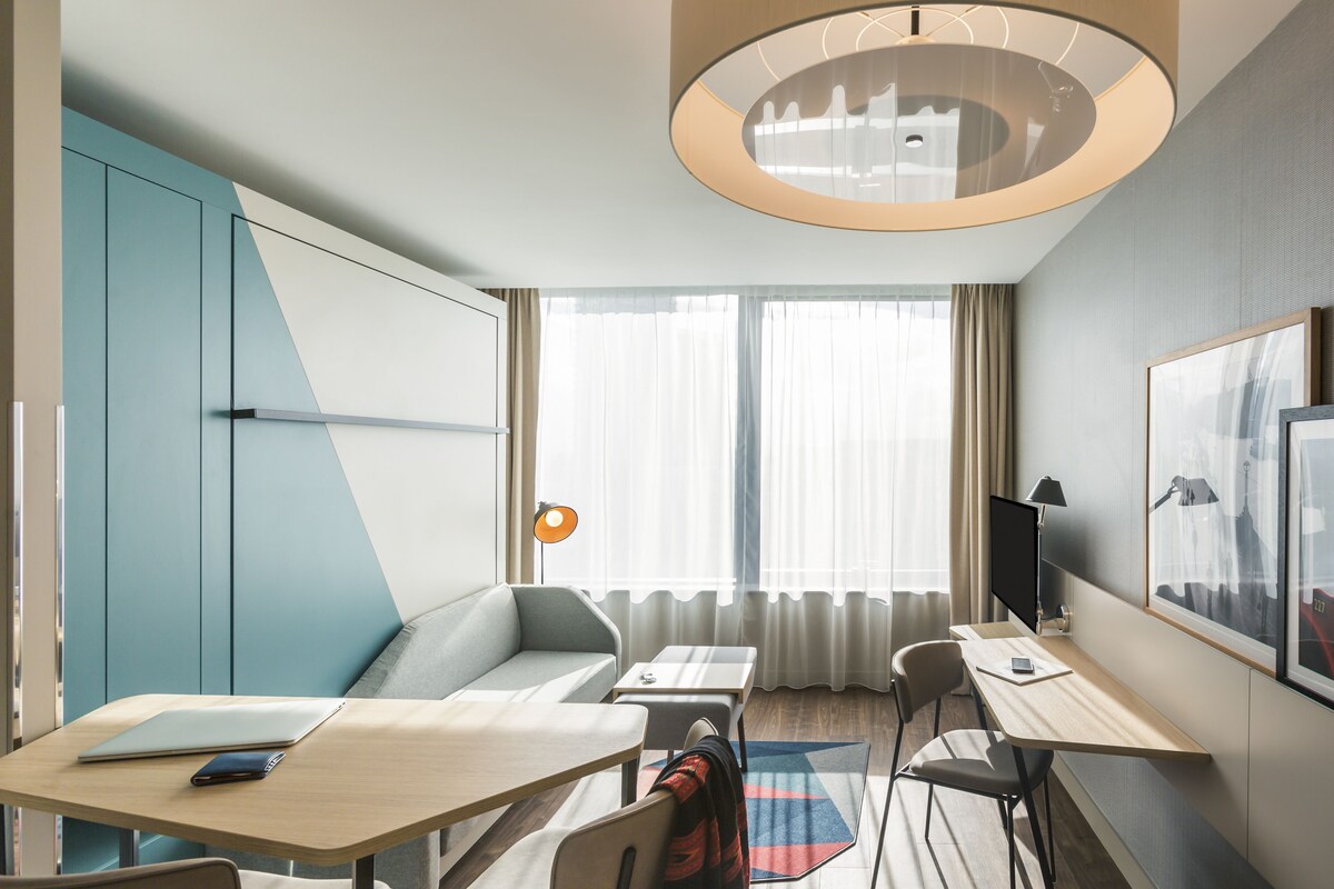 Una habitación de hotel moderna con un sofá, un escritorio, una computadora portátil, un teléfono inteligente y un televisor.
