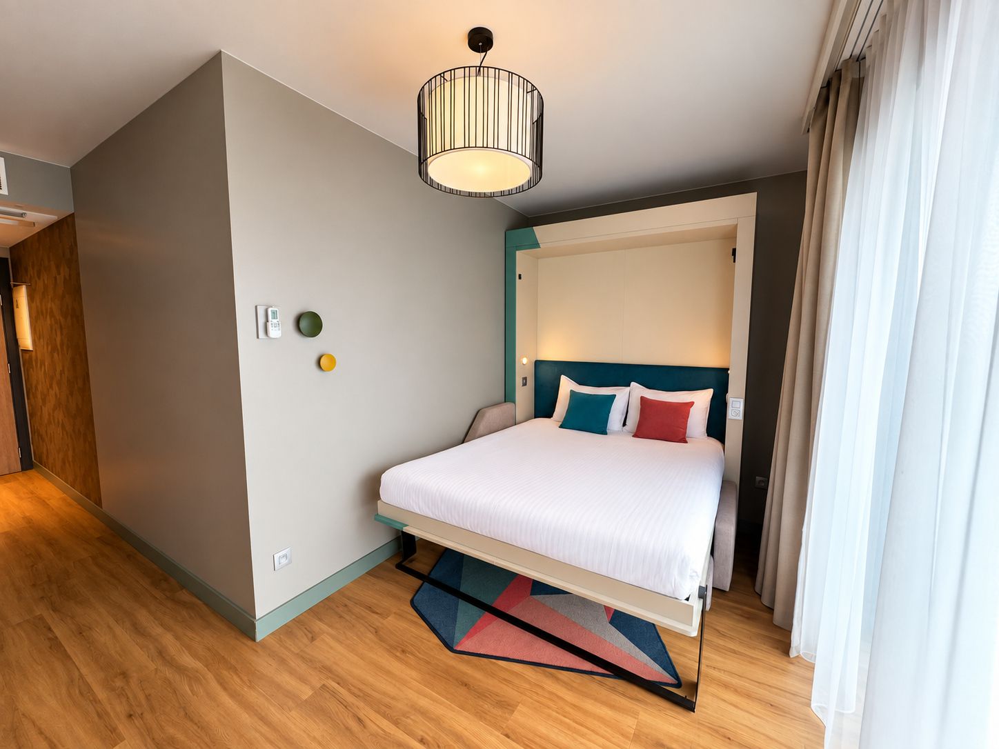 A modern hotel room with a bed, a ceiling light, a wall-mounted thermostat, and a window with curtains.