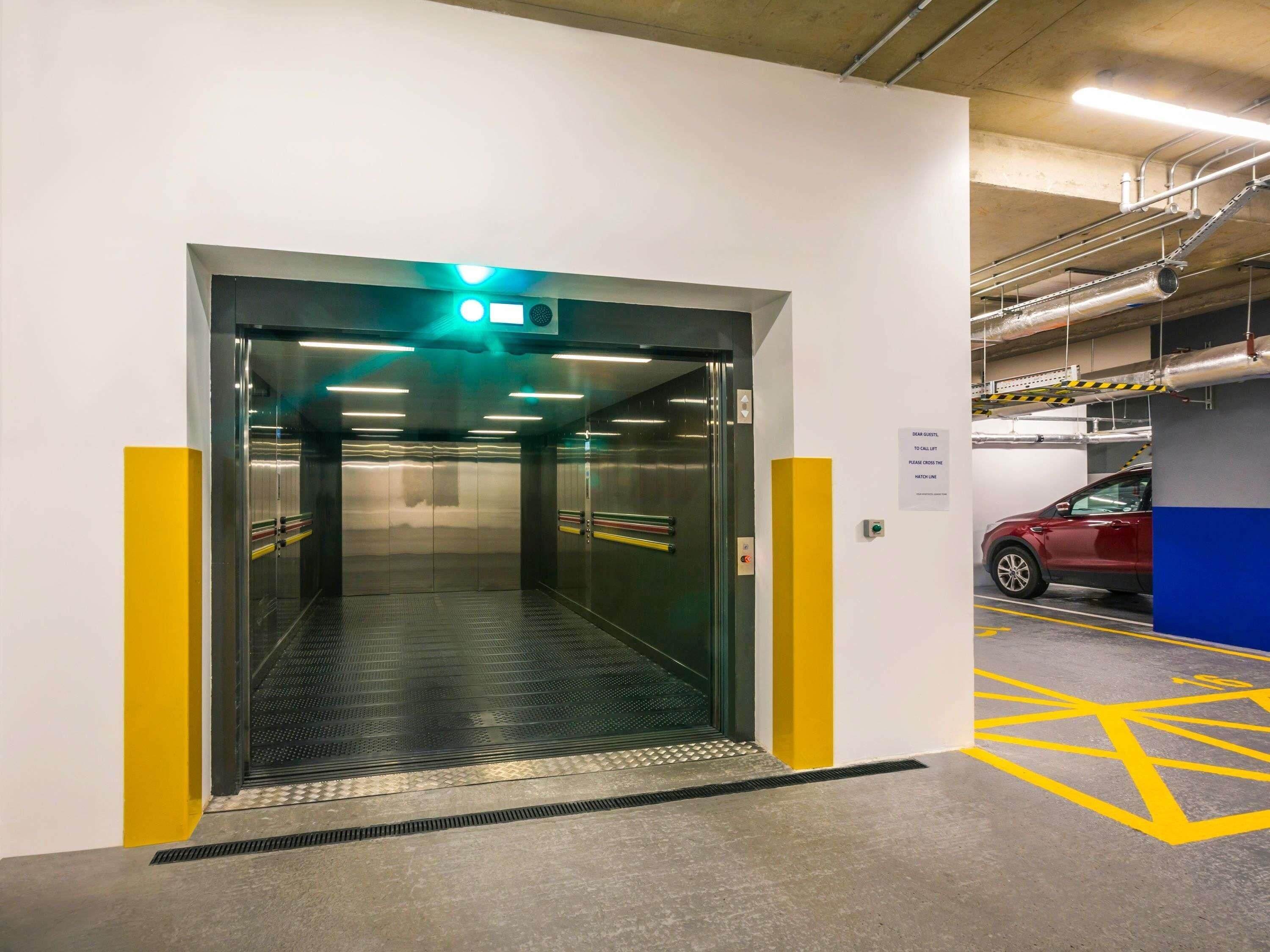 En hiss med en grön utrymningsskylt, en röd bil parkerad i ett garage och gula parkeringslinjer på golvet.