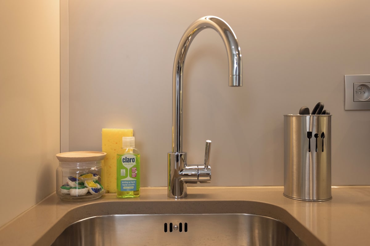 Un lavamanos de acero inoxidable con un grifo moderno, un tarro de utensilios de cocina, una botella de detergente y un recipiente con utensilios.