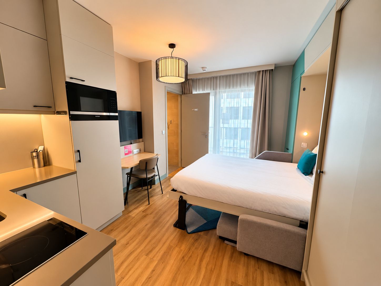 Habitación de hotel compacta con kitchenette, una cama con cabecera, un escritorio con silla y un ventanal con cortinas.