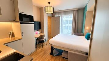 Habitación de hotel compacta con kitchenette, una cama con cabecera, un escritorio con silla y un ventanal con cortinas.