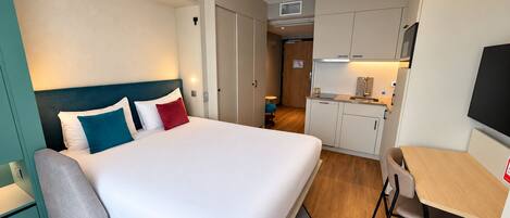 A hotel room with a bed, a desk, a chair, a kitchenette, and a television.