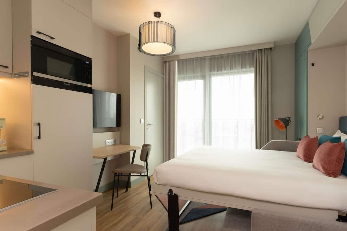 Quarto de hotel moderno com cama, escrivaninha, cadeira, televisão e janela com cortinas.
