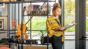 A person reading a book in a room with a bookshelf containing various items, a guitar, and a sign that reads "LIBRARY". - Adagio Antwerp City Center (Antwerp)