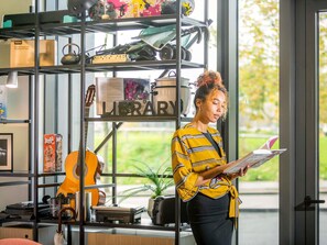 Una persona leyendo un libro en una habitación con una estantería que contiene diversos objetos, una guitarra y un letrero que dice “LIBRARY”.