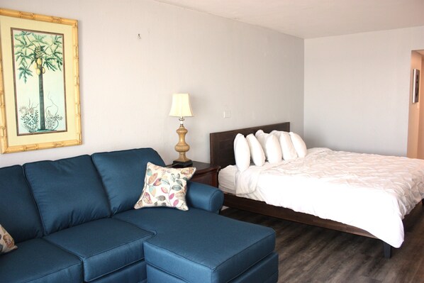 A room with a blue sofa, a bed with white bedding, a lamp, and a framed picture on the wall.