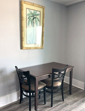 A dining area with a table and two chairs, a framed picture on the wall, and a light-colored wall.