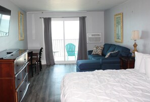 A hotel room with a bed, a desk with a chair, a blue sofa, a balcony with a chair, and a lamp.