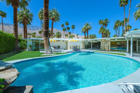 A pool area with a clear blue pool, surrounded by palm trees and a modern building with outdoor seating. - Midcentury Masterpiece in Deepwell Estates - Pool and View  (Palm Springs)