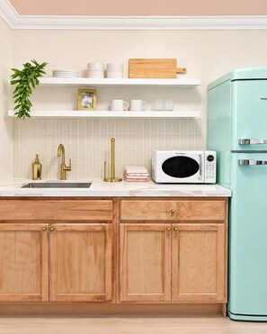 Une cuisine avec des meubles en bois, un plan de travail blanc, un évier avec un robinet doré, un micro-ondes blanc et un réfrigérateur de couleur turquoise.