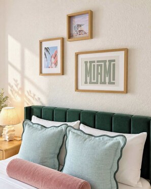 Un lit avec une tête de lit recouverte d’un tissu vert, deux oreillers, une lampe de chevet et des tableaux encadrés accrochés au mur.
