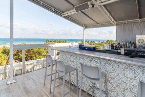 A covered outdoor bar area with a view of the ocean, bar stools, and a variety of bottles.