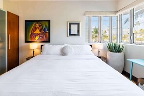 A hotel room with a large bed, a painting on the wall, a potted plant, and a view of palm trees through the windows. - Oceanfront Oasis: 2-BR Apartment on Ocean Drive (Miami Beach)