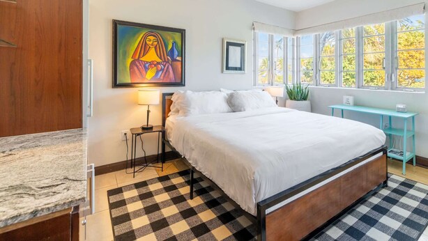 Een slaapkamer met een groot bed, een zwart-wit schaakbordvloer, een schilderij aan de muur en een raam met uitzicht op bomen.