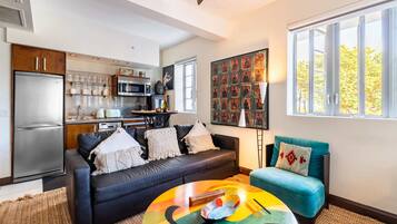 Un salon moderne avec un canapé noir, une table basse colorée et un fauteuil turquoise. On aperçoit, au fond, une cuisine équipée d’un réfrigérateur et d’un four à micro-ondes.