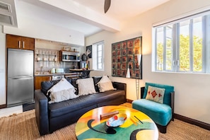 Un salon moderne avec un canapé noir, une table basse colorée et un fauteuil turquoise. On aperçoit, au fond, une cuisine équipée d’un réfrigérateur et d’un four à micro-ondes.
