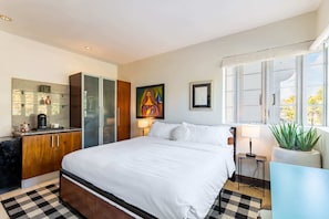 Une chambre d’hôtel avec un grand lit, une kitchenette, un tableau, une fenêtre donnant sur un beau paysage et une plante en pot.