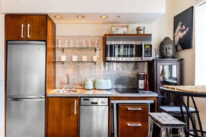A modern kitchen with stainless steel appliances, wooden cabinets, a glass shelf with cups, a microwave, and a Buddha statue. - Oceanfront Oasis: 2-BR Apartment on Ocean Drive (Miami Beach)