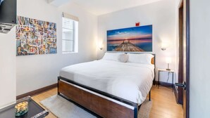 A neatly arranged bedroom with a large bed, a small bedside table, a wall-mounted TV, and a painting of a sunset over a pier. - Oceanfront Oasis: 2-BR Apartment on Ocean Drive (Miami Beach)