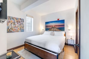 Une chambre à coucher bien rangée, avec un grand lit, une petite table de chevet, un téléviseur fixé au mur et un tableau représentant un coucher de soleil sur une jetée.