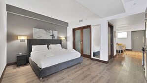 Una habitación de hotel moderna con una cama grande, lámparas de noche, piso de madera y vista al comedor.